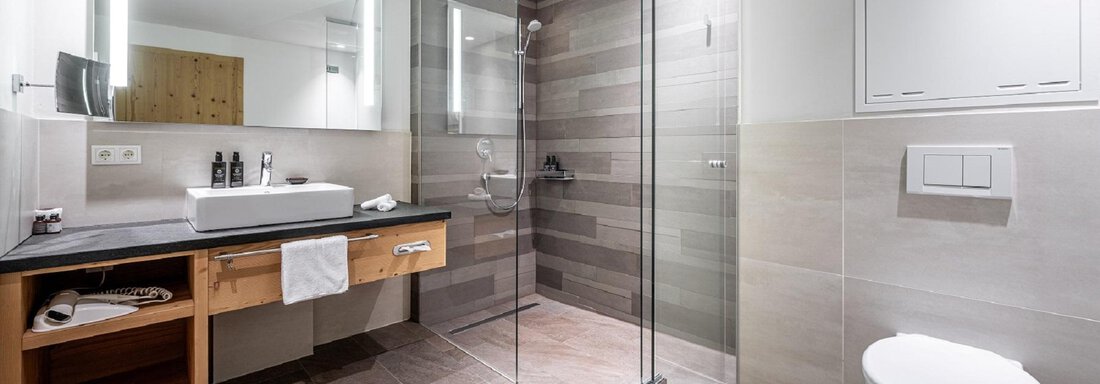 A modern bathroom with a glass shower and a wooden vanity. The walls are painted in neutral colors, and there is ample light from the mirror lighting. - Gletscherblick - Serviced Apartments - Obergurgl 