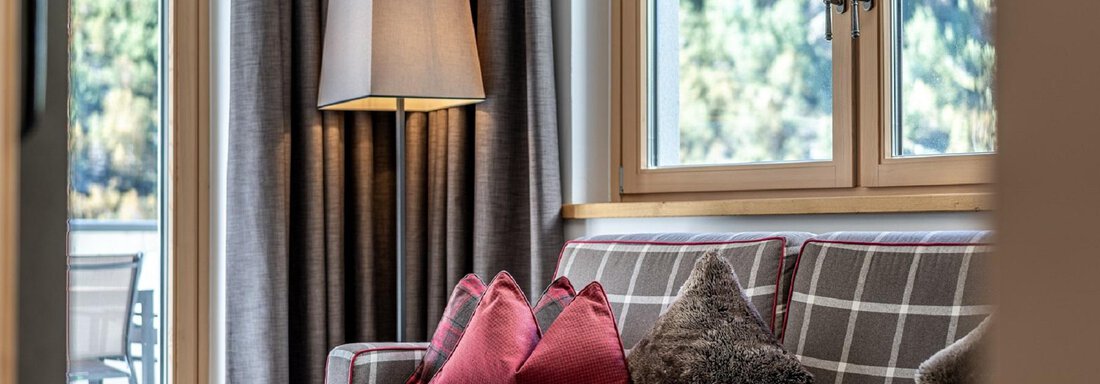 A cozy living room with a sofa and cushions in warm colors. Large windows provide plenty of daylight and an inviting atmosphere. - Gletscherblick - Serviced Apartments - Obergurgl 