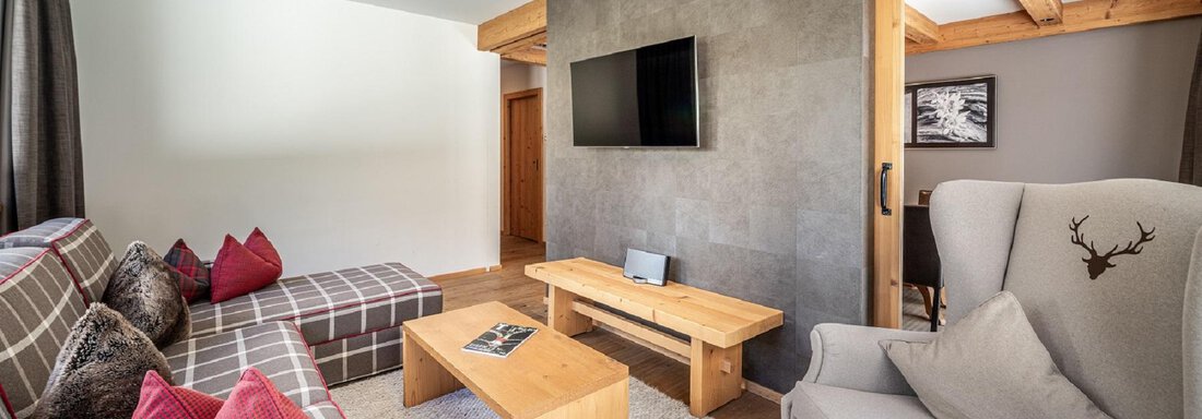 A cozy living room with modern furniture and warm colors. There is a large window, a television, and comfortable seating. - Gletscherblick - Serviced Apartments - Obergurgl 