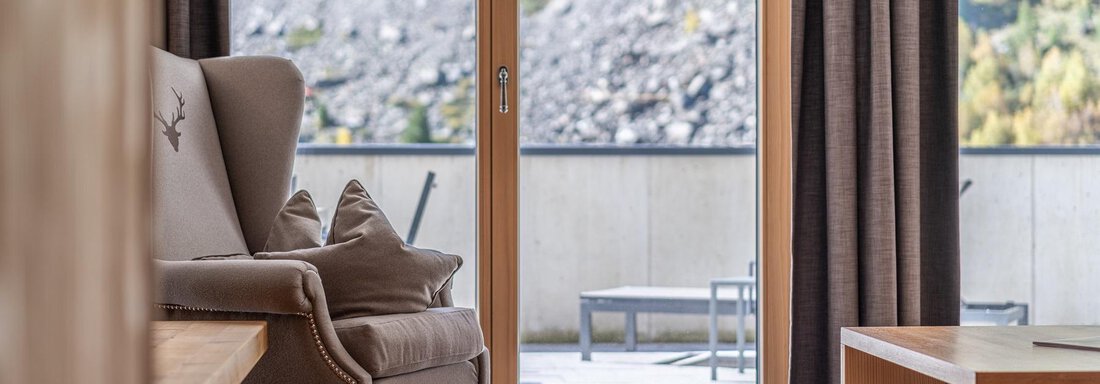 Apt_2_Wohnen A cozy armchair is in a modern room with large windows. View of nature and a terrace with chairs. - Gletscherblick - Serviced Apartments - Obergurgl