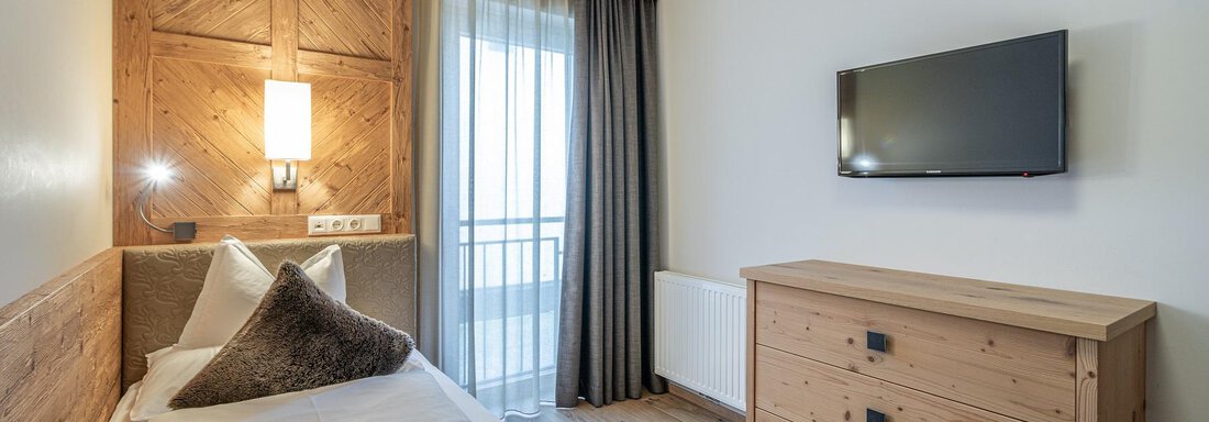 A modern hotel room with wooden paneling and daylight. It features a bed, a television, and a dresser. - Gletscherblick - Serviced Apartments - Obergurgl 