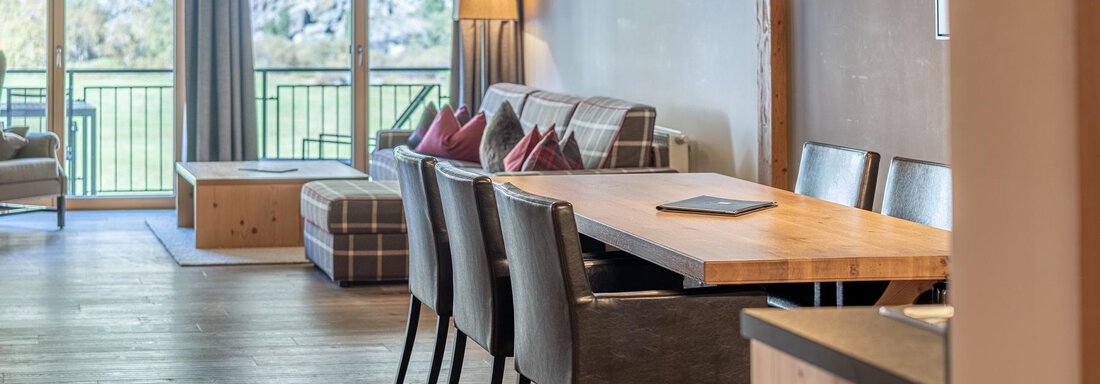 A modern living room with a large dining table and black chairs. Large windows offer a view of nature. - Gletscherblick - Serviced Apartments - Obergurgl 