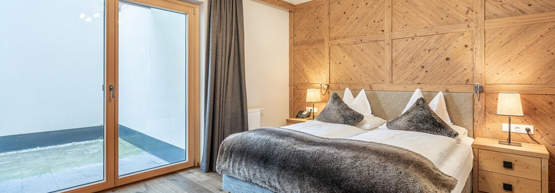A modern bedroom with wooden paneling and a large window. The room is elegantly furnished and has a cozy atmosphere. - Gletscherblick - Serviced Apartments - Obergurgl 