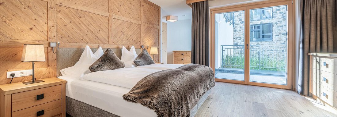 A modern bedroom with a double bed, wood paneling, and elegant lighting. Large windows offer a view of the nature outside. - Gletscherblick - Serviced Apartments - Obergurgl 