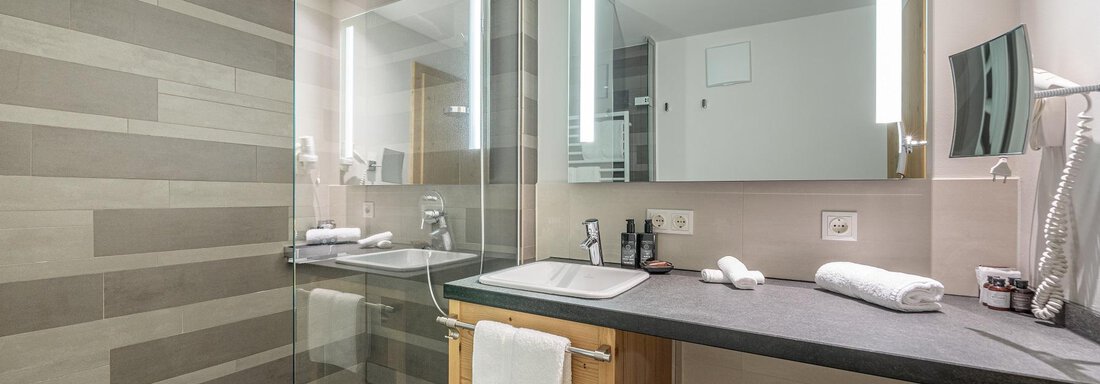 A modern bathroom with a glass shower cabin and a simple washbasin. The walls are in neutral colors and there is mirror lighting. - Gletscherblick - Serviced Apartments - Obergurgl 