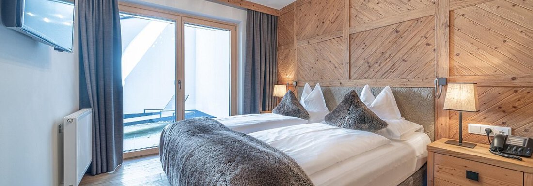 A bright bedroom with a large double bed and elegant wooden paneling. Large windows provide access to a balcony and natural light. - Gletscherblick - Serviced Apartments - Obergurgl 