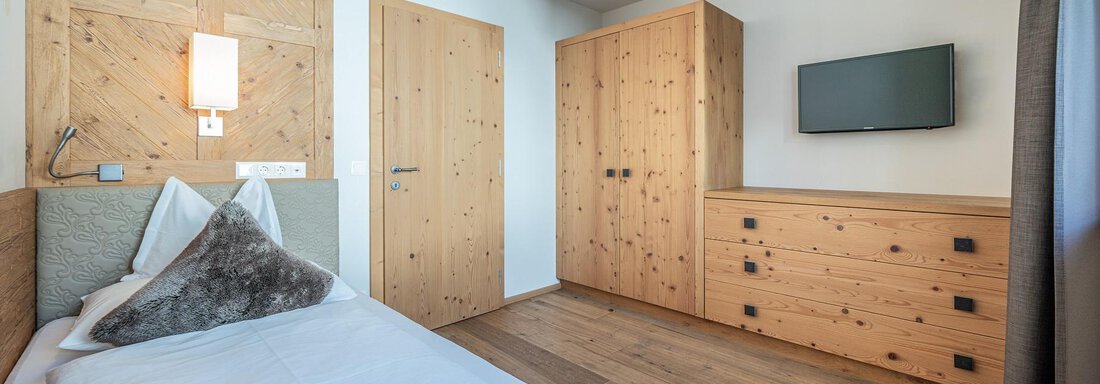 A bright room with wooden walls and a single bed. There is a dresser, a television, and a window with curtains. - Gletscherblick - Serviced Apartments - Obergurgl 
