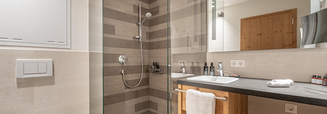 A modern bathroom with a glass shower and a stylish wooden shelf. The walls are in soft earth tones. - Gletscherblick - Serviced Apartments - Obergurgl 