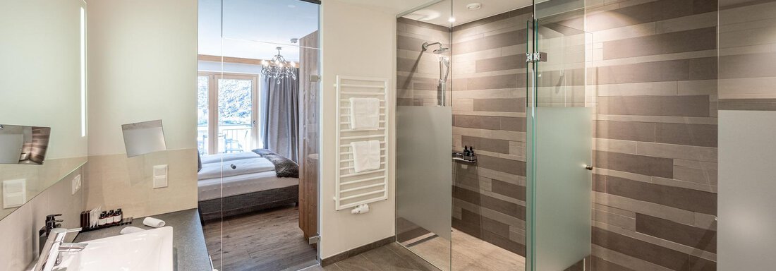 A modern bathroom with a glass shower and a large sink. In the background, a cozy bedroom is visible. - Gletscherblick - Serviced Apartments - Obergurgl 