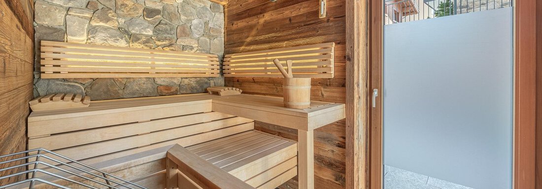 A cozy sauna with wooden interior and a stone area. The door offers a view outside and lets light in. - Gletscherblick - Serviced Apartments - Obergurgl 