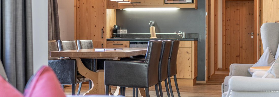 A modern living room with a stylish kitchen in the background. The dining table is decorated with black chairs, and the walls are in warm wood tones. - Gletscherblick - Serviced Apartments - Obergurgl 
