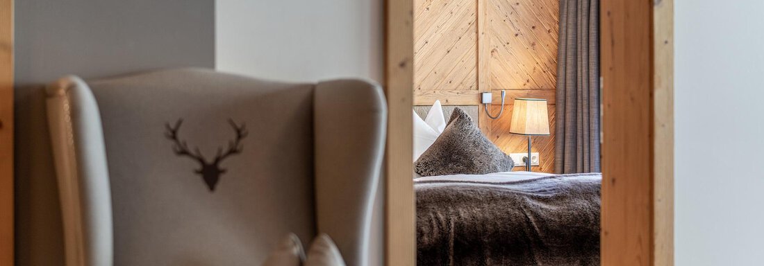 A cozy room with a comfortable armchair and a chandelier. In the background, a bedroom with wood paneling and a warm atmosphere is visible. - Gletscherblick - Serviced Apartments - Obergurgl 