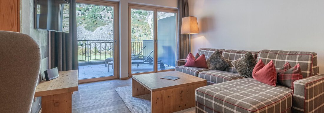 A cozy living room with a large, comfortable sofa and a wooden table. Large windows offer a view of the outdoor area. - Gletscherblick - Serviced Apartments - Obergurgl 