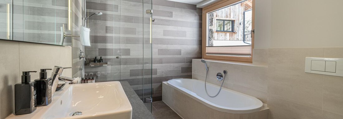 A modern bathroom with a bathtub and a separate shower. Large windows provide plenty of natural light. - Gletscherblick - Serviced Apartments - Obergurgl 