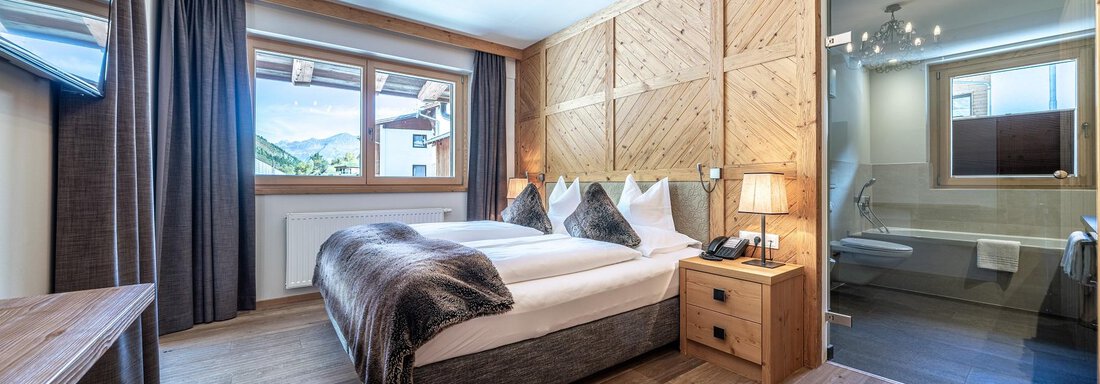 A modern hotel room with a double bed and wooden paneling. The room has large windows that offer a view of the landscape. - Gletscherblick - Serviced Apartments - Obergurgl 