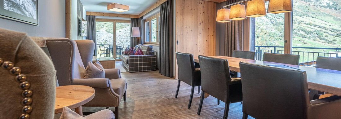 A stylish living room with wooden walls and large windows that offer a view of the mountains. The room is furnished with cozy furniture and a dining area. - Gletscherblick - Serviced Apartments - Obergurgl 