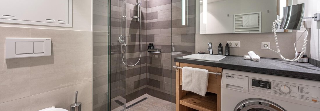 A modern bathroom with a glass shower and a washing machine. The walls are designed in neutral tones and there is a bright mirror. - Gletscherblick - Serviced Apartments - Obergurgl 