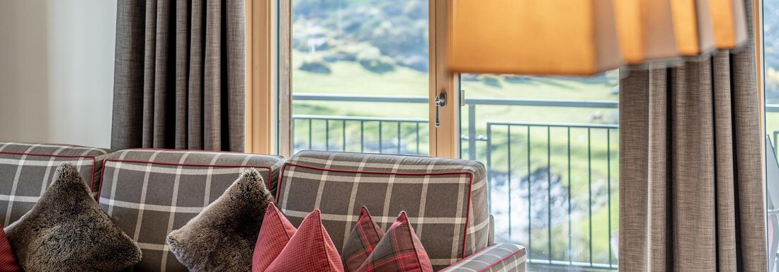 A cozy living room with a checkered sofa and colorful cushions. Large windows offer a beautiful view of the landscape. - Gletscherblick - Serviced Apartments - Obergurgl 