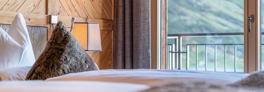 Apt_14_Detail A cozy bedroom with a large window overlooking a green landscape. The bedding is light, and the room has wooden details for a warm atmosphere. - Gletscherblick - Serviced Apartments - Obergurgl
