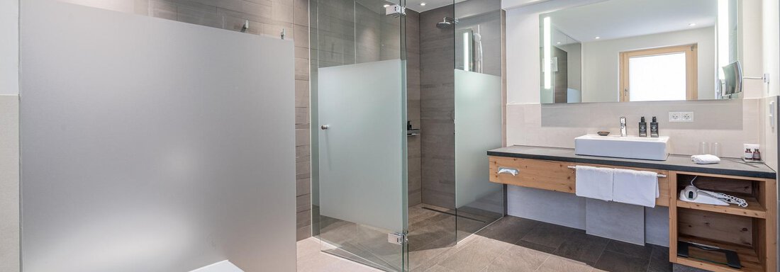 A modern bathroom with a glass shower and a large bathtub. The space is bright and elegantly designed, with a wooden vanity and stylish fixtures. - Gletscherblick - Serviced Apartments - Obergurgl 