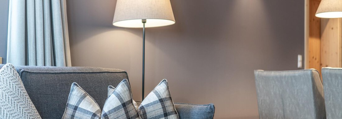 A modern living room with a gray sofa and decorative cushions. A floor lamp and a dining table in the background complement the cozy space. - Gletscherblick - Serviced Apartments - Obergurgl 