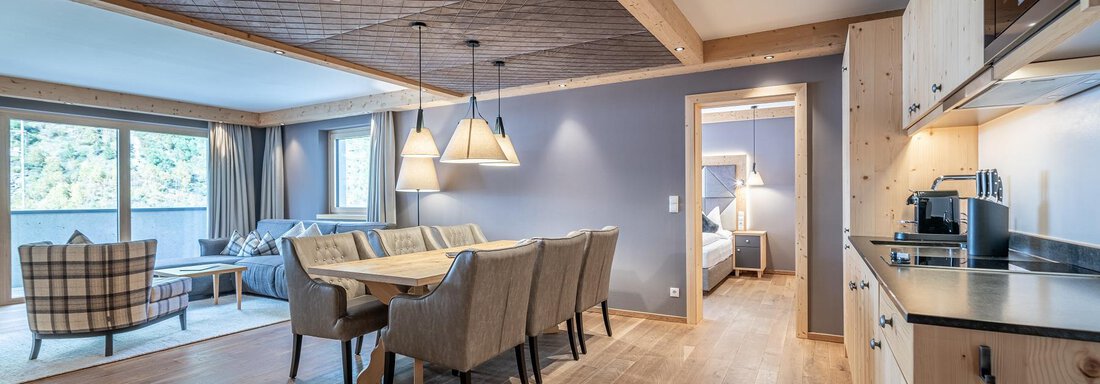 An elegant living area with a large dining table and cozy chairs. The wooden ceiling and warm floor give the room an inviting atmosphere. - Gletscherblick - Serviced Apartments - Obergurgl 