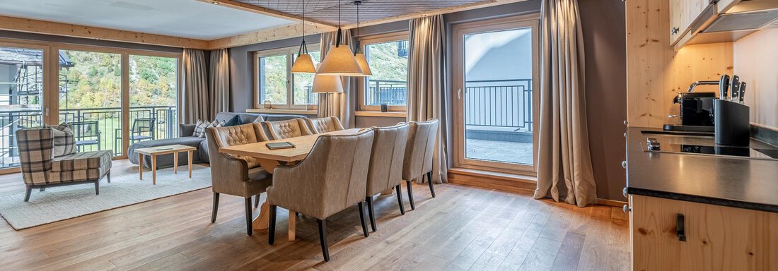 A modern living room with a large dining table and comfortable chairs. Large windows provide plenty of daylight and a view of the balcony. - Gletscherblick - Serviced Apartments - Obergurgl 