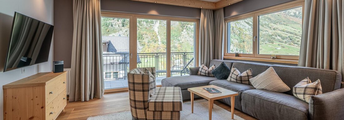 Apt_23_Wohnen A modern living room with large windows and a view of the mountains. The decor is cozy with a sofa, an armchair, and a wooden table. - Gletscherblick - Serviced Apartments - Obergurgl