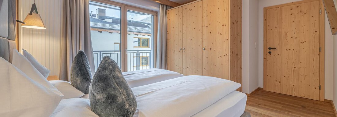 A modern bedroom with two beds, natural wood paneling, and soft lighting. Large windows provide plenty of daylight and an inviting atmosphere. - Gletscherblick - Serviced Apartments - Obergurgl 