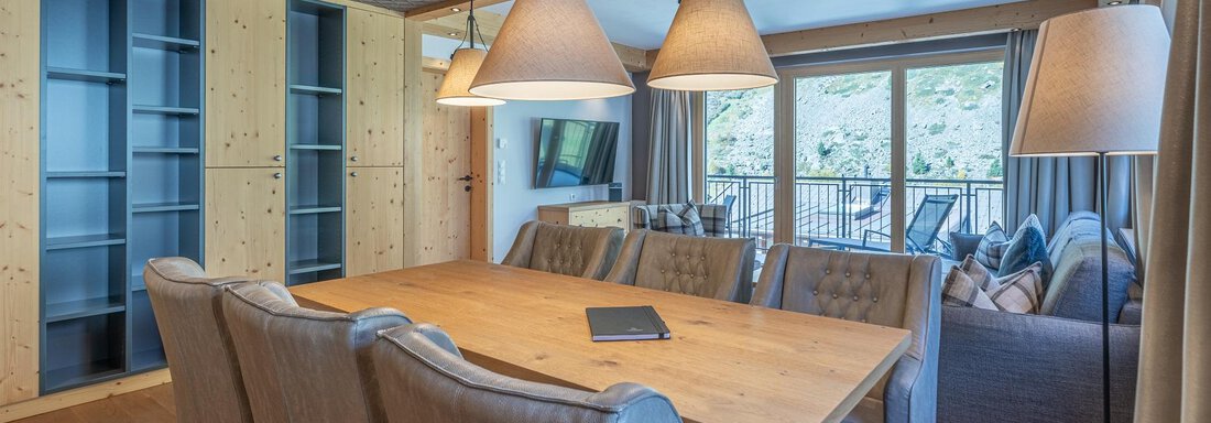 A modern dining room with a large wooden table and elegant chairs. Large windows offer a view of the outdoor terrace and let in plenty of light. - Gletscherblick - Serviced Apartments - Obergurgl 