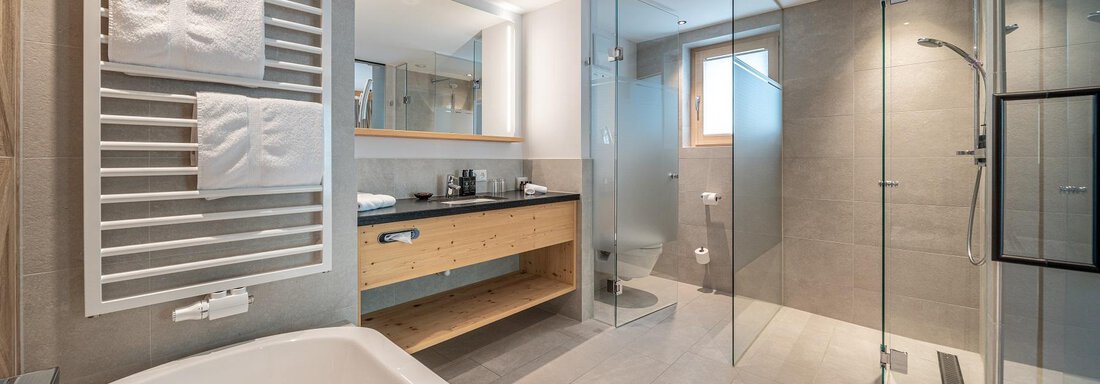 A modern bathroom with a contemporary bathtub and a separate shower. Bright tiles and wooden shelves create an inviting atmosphere. - Gletscherblick - Serviced Apartments - Obergurgl 