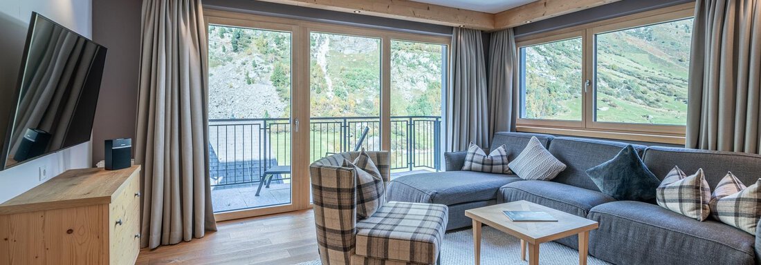 A modern living room with large windows and a view of the mountains. There is a cozy sofa and an armchair, as well as a wooden furniture chest. - Gletscherblick - Serviced Apartments - Obergurgl 