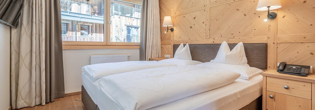 A cozy hotel room with a large bed and wooden paneling. There are natural light sources and a peaceful atmosphere. - Gletscherblick - Serviced Apartments - Obergurgl 