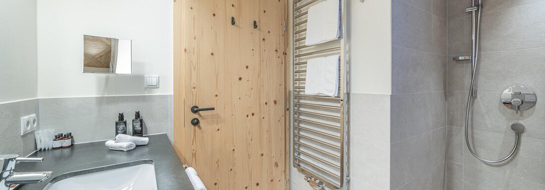 A modern bathroom with a shower and a sink. The walls are light, and the door is made of wood. - Gletscherblick - Serviced Apartments - Obergurgl 