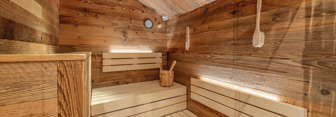 A cozy sauna with wooden paneling on the walls and benches. In the room, there is a bucket and a spade. - Gletscherblick - Serviced Apartments - Obergurgl 