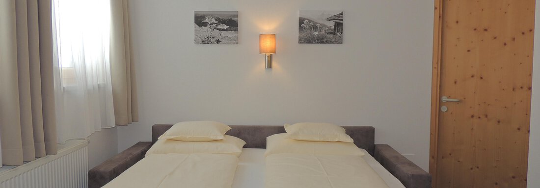 A simple bedroom with a double bed and two pillows. The walls are light and there are two pictures as well as a wooden shelf. - Haus Christophorus - Obergurgl 