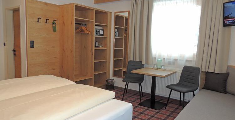 A modern hotel room with two beds, a sofa, and a small table. The room has a large window with curtains and wooden shelves. - Haus Christophorus - Obergurgl 