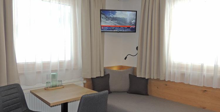A cozy living space with a sofa, a table, and two chairs. A television hangs on the wall, and the windows are adorned with light curtains. - Haus Christophorus - Obergurgl 
