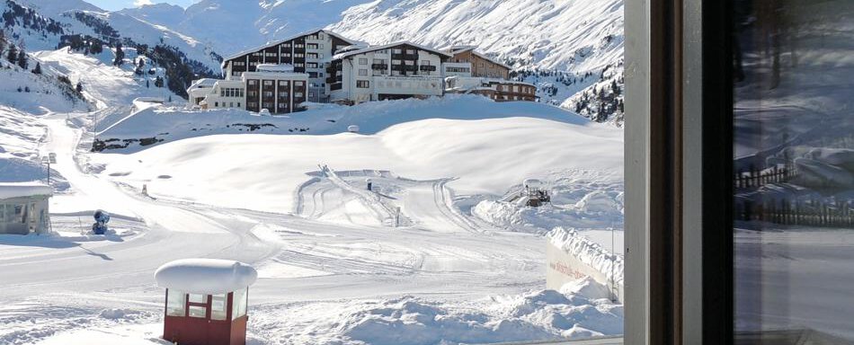 A snowy landscape with a building in the background and mountains. The sunlit scene conveys a peaceful winter atmosphere. - Haus Christophorus - Obergurgl 