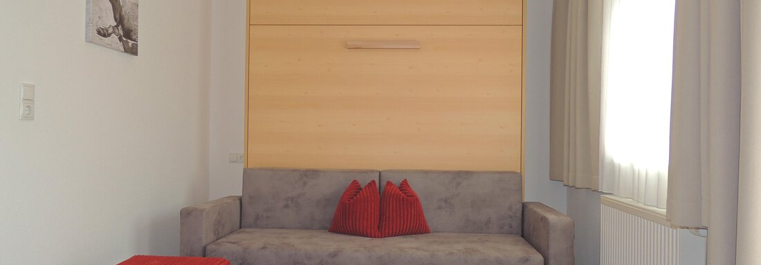 A modern living room with a gray sofa and two red cushions. A small wooden table stands in front of the sofa, and there is a red pouf as well as large windows with curtains. - Haus Christophorus - Obergurgl 