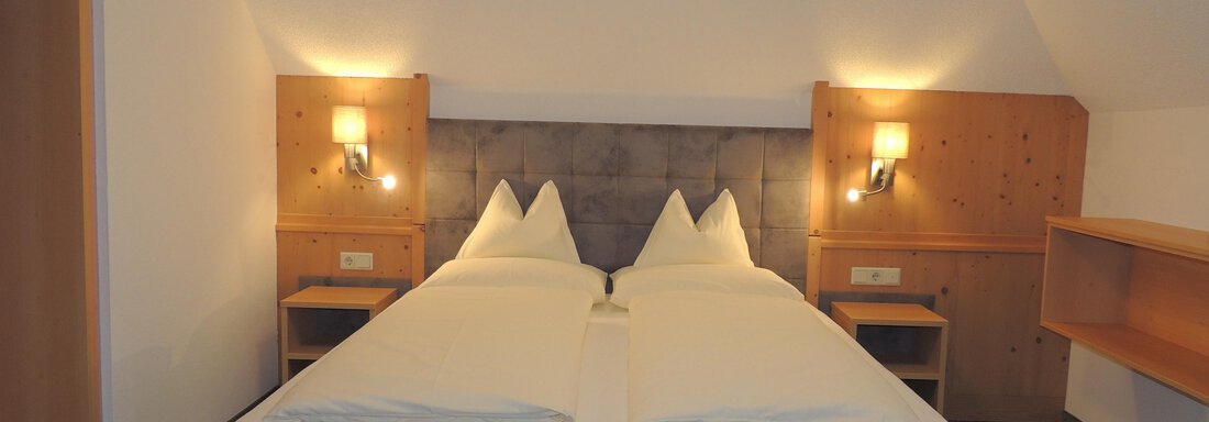 A cozy hotel room with a double bed and bright walls. There are two bedside tables and a wooden shelf on the wall. - Haus Christophorus - Obergurgl 
