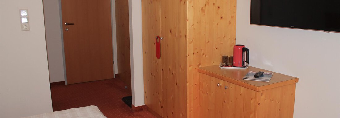 A simple room with two beds, a wooden wardrobe, and a television on the wall. On a table, there is a kettle and some utensils. - Haus Martinshof - Obergurgl 