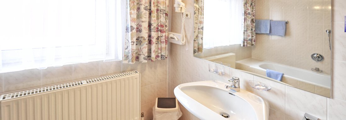 A modern bathroom with a bathtub, a sink, and a large window. The walls are light, and the windows are adorned with curtains. - Haus Timmelseck - Obergurgl 