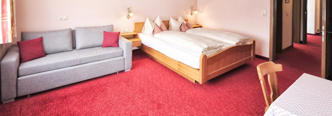 A cozy hotel room with a red carpet, a double bed, and a gray sofa bed. A white tablecloth is on the table, and the walls are decorated with pictures. - Haus Timmelseck - Obergurgl 