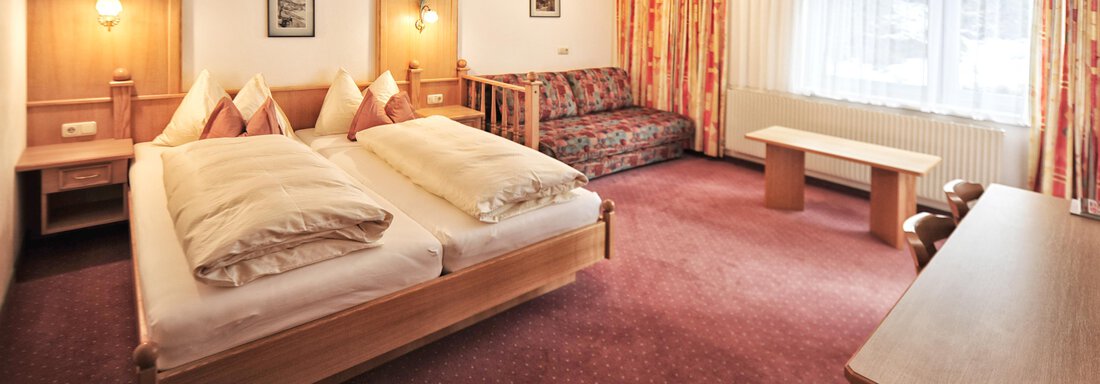 A cozy hotel room with two beds, a sofa, and a table. Large windows provide plenty of daylight. - Haus Timmelseck - Obergurgl 