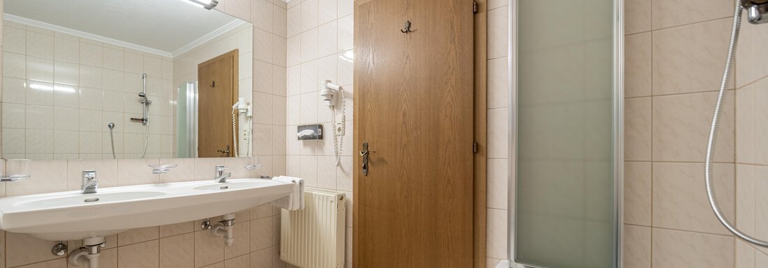 A simple bathroom with a bathtub, a large mirror, and a wooden or plastic door. The walls are tiled in a light color and the floor is in a neutral shade. - Haus Timmelseck - Obergurgl 