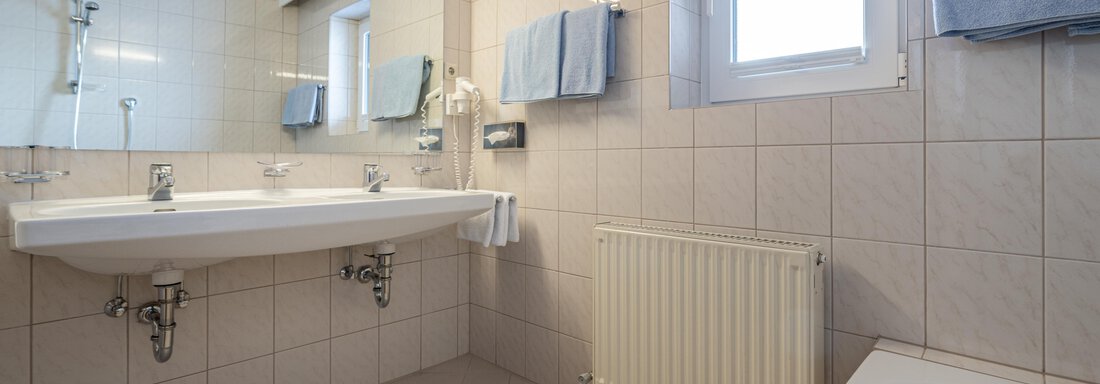 A modern bathroom with a large sink area and a window. The walls are light tiled and there are fresh blue towels. - Haus Timmelseck - Obergurgl 