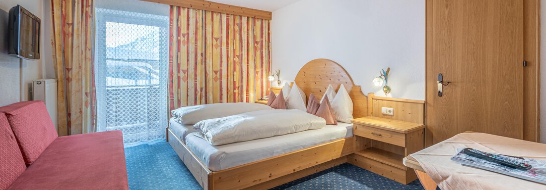 A cozy bedroom with two beds and a red sofa. There is a balcony with a view outside and a table with chairs. - Haus Timmelseck - Obergurgl 