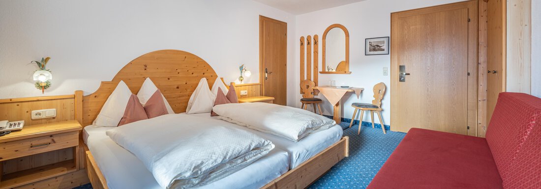 A cozy hotel room with a double bed and red accents. There is a desk and a red sofa, as well as warm wood decor. - Haus Timmelseck - Obergurgl 