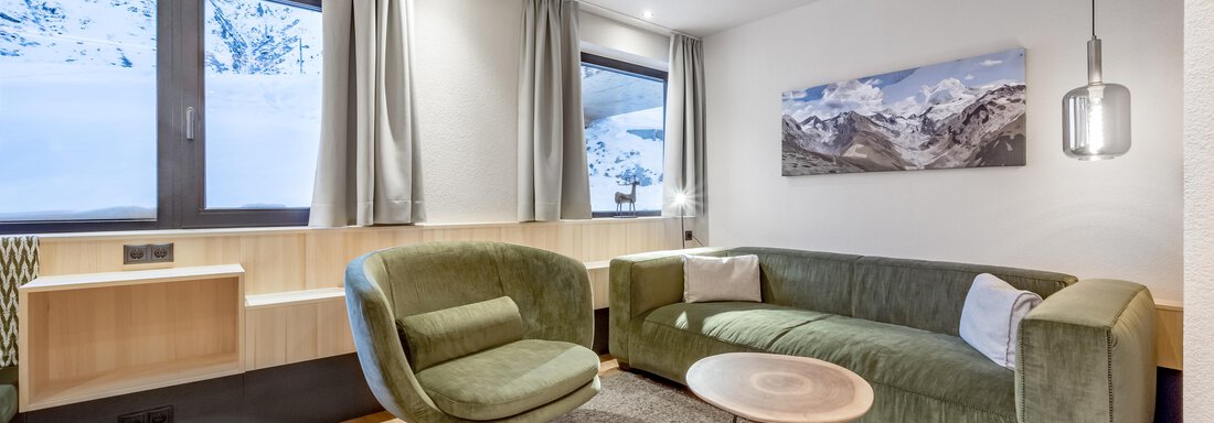 Apartement Alpenglück XL de luxe A modern living room with a cozy green sofa and a round table. Large windows offer a view of the snowy mountains. - Hotel-Garni Pirchhütt - Obergurgl